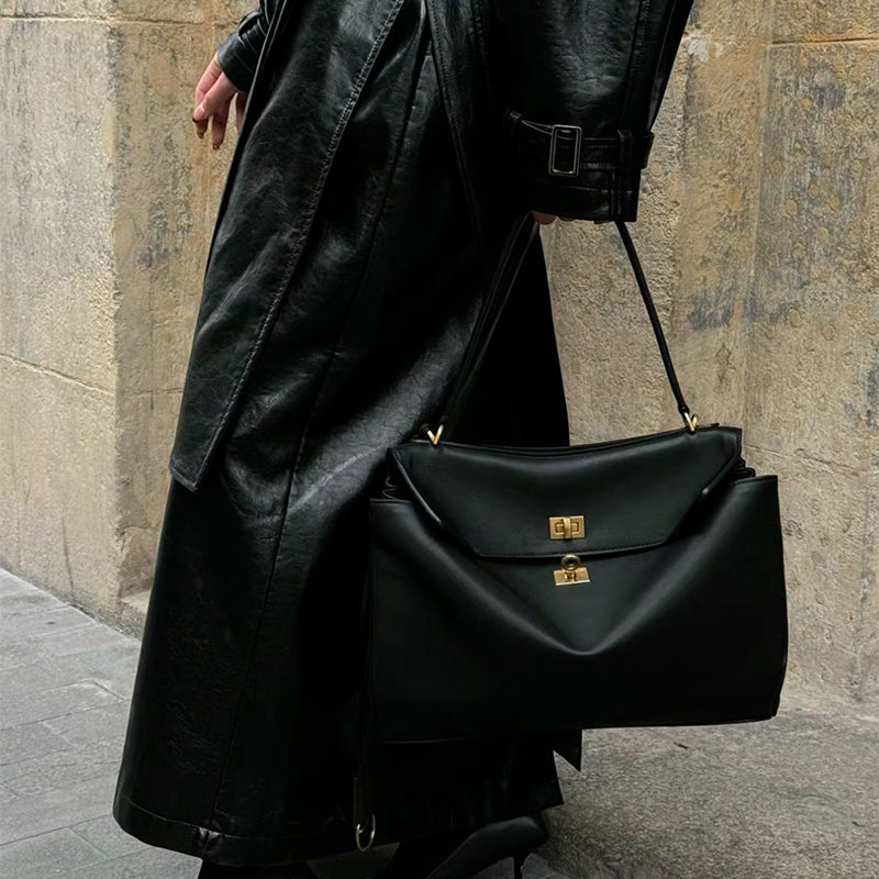 Person wearing a black coat and carrying a black handbag with gold accents against a stone wall.