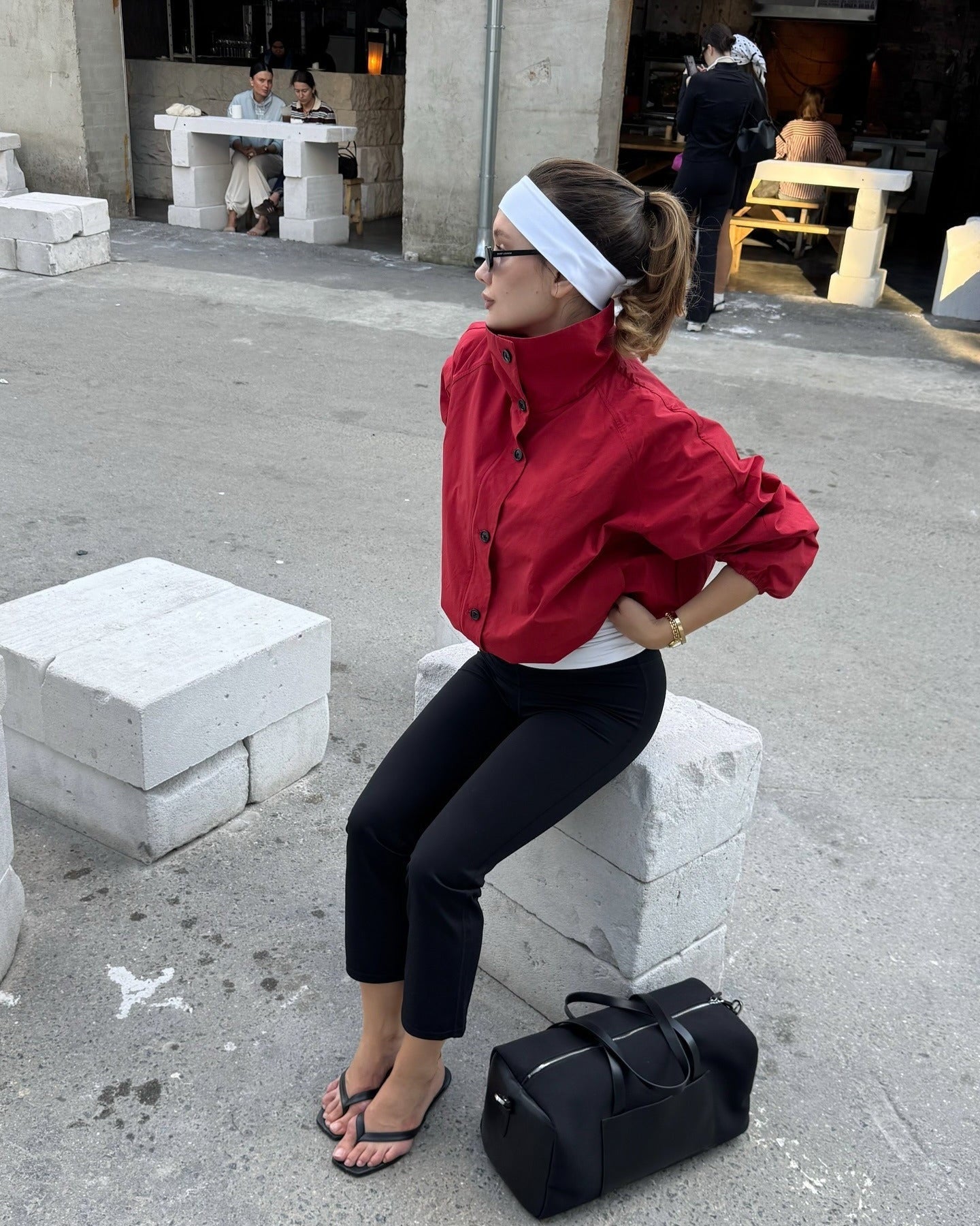 Woman in red jacket and black pants sitting on a concrete block with a black duffel bag beside her.